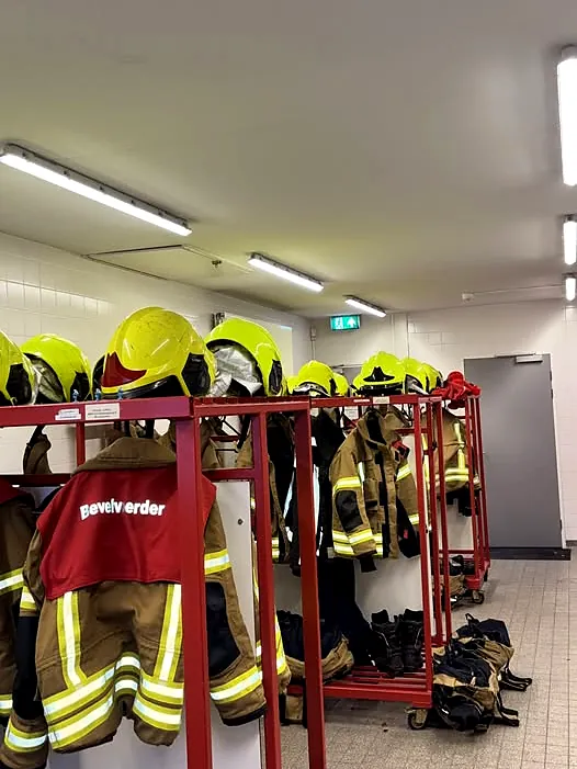 Kleedruimte van de kazerne met uitrukkleding aan de rekken, verlicht door TL-armaturen aan het plafond